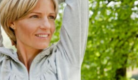A woman with short blonde hair smiling during a stretch outdoors, enjoying a healthy lifestyle surrounded by greenery in Los Angeles.