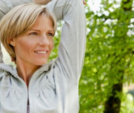 A woman with short blonde hair smiling during a stretch outdoors, enjoying a healthy lifestyle surrounded by greenery in Los Angeles.
