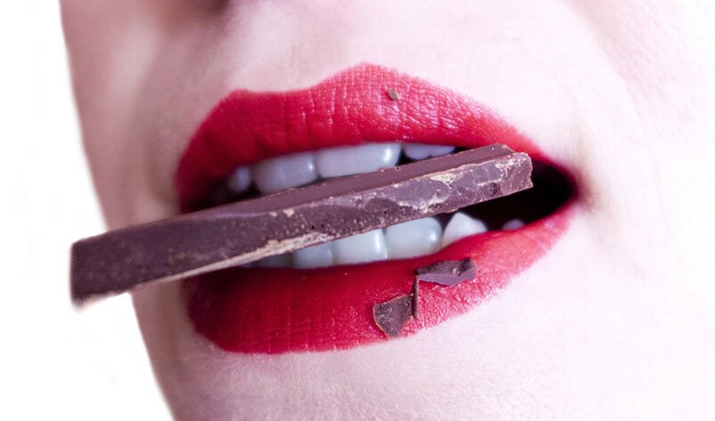 Indulging in a sweet escape: a close-up of vibrant red lips biting into a rich piece of dark chocolate, recommended by a sexual health specialist in Los Angeles.