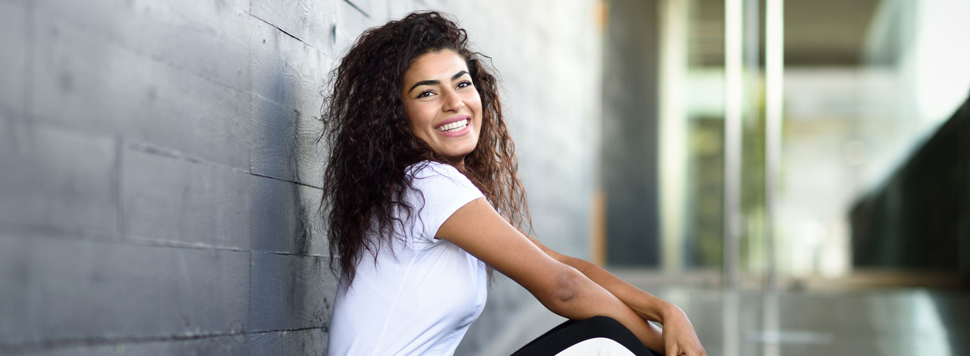 A smiling woman with curly hair, who is a sexual health specialist in Los Angeles, sitting casually against a gray wall in a modern, airy space.