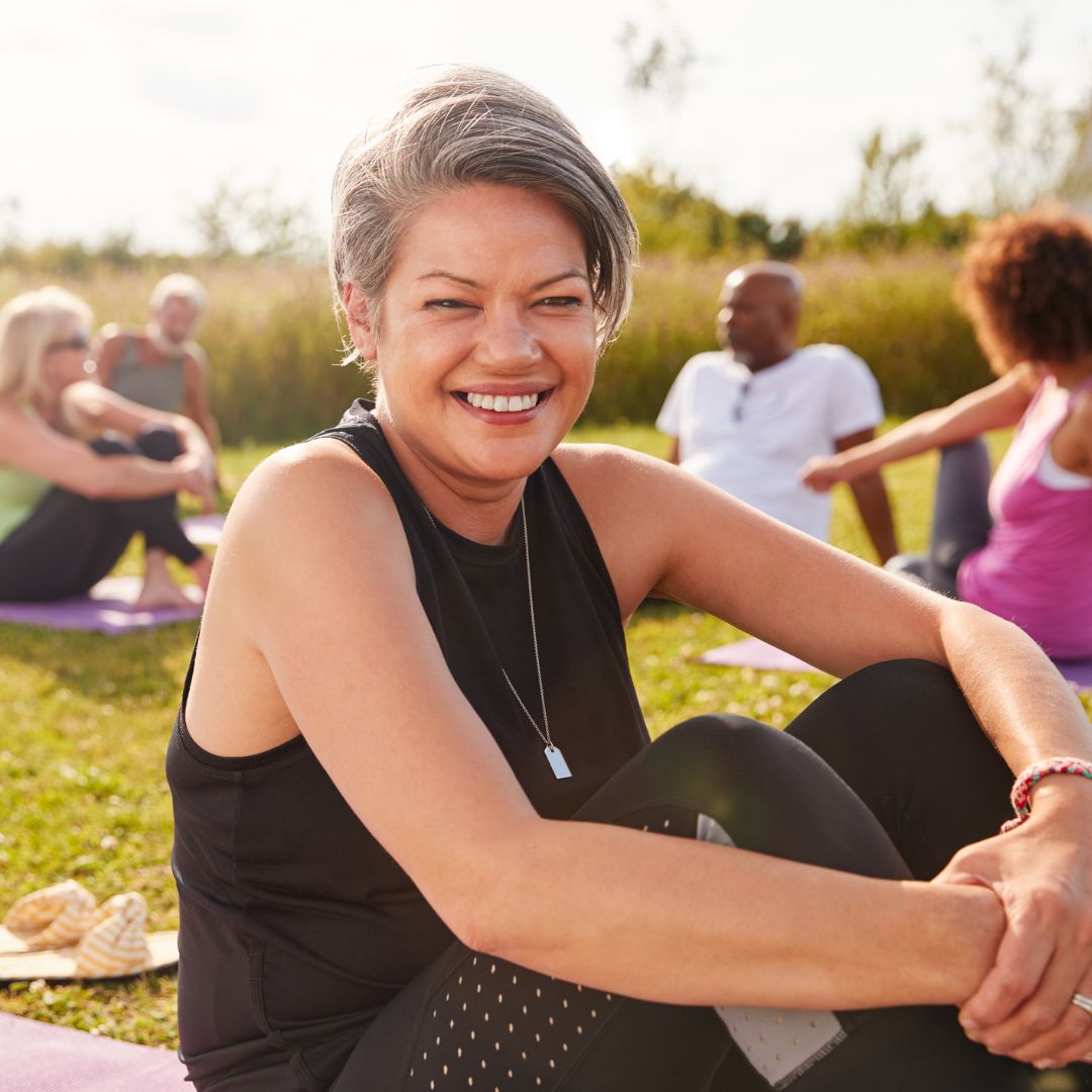 A smiling woman enjoying a relaxing moment at an outdoor yoga session with a group of diverse people in the background, led by a renowned sexual health specialist from Los Angeles.