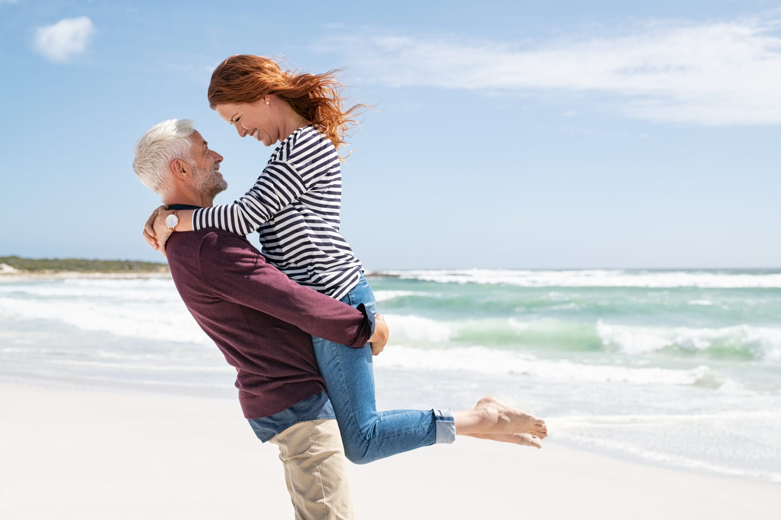 A happy couple sharing a joyful moment as a man lifts a woman in a warm embrace on a sunny beach, with gentle waves in the background, after consulting with their sexual health specialist in Los Angeles.