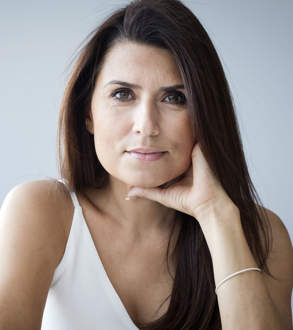 A confident woman with long dark hair and a white top gazes intently at the camera, resting her chin on her hand, embodying the transformative results of consulting a sexual health specialist in Los Angeles