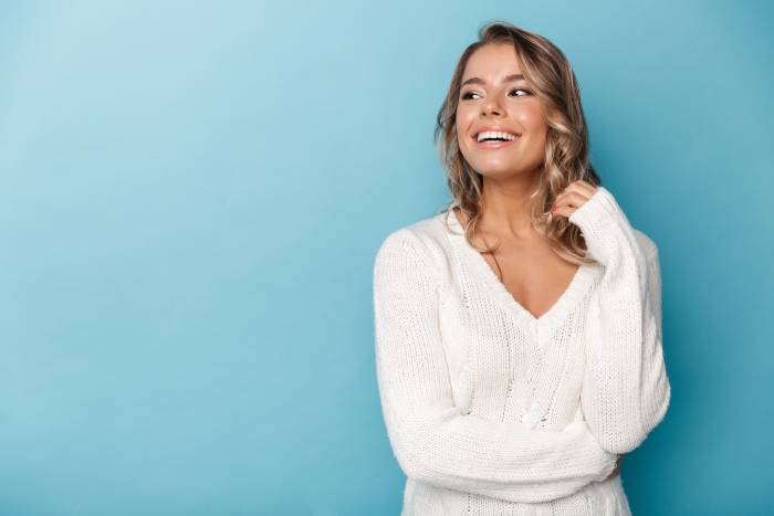 A cheerful young woman in a white sweater, joyfully looking away on a light blue background, contemplates visiting a sexual health specialist in Los Angeles.
