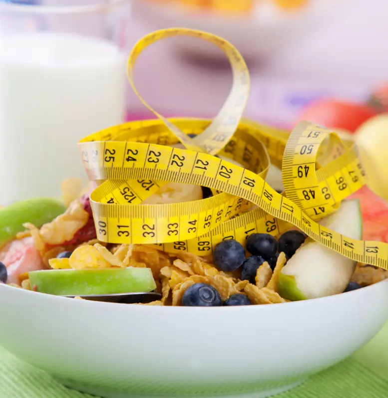 A bowl of healthy breakfast cereal with fruits wrapped in a measuring tape, symbolizing a nutritious diet and portion control for weight management as advised by a Sexual Health Expert in Los Angeles.