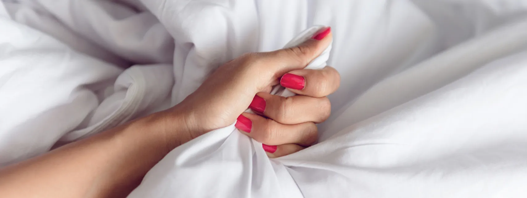 A close-up of a person's hands with pink nail polish, clutching a white fabric, possibly a bedsheet, suggesting a moment of emotion or tension discussed during a consultation with a Sexual Health Expert