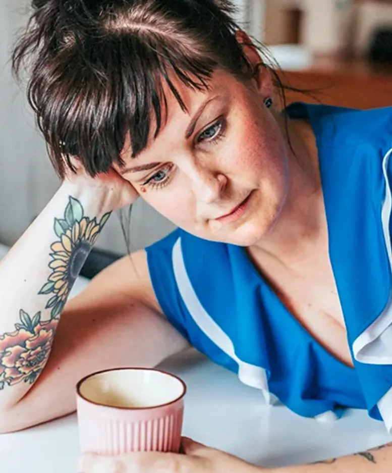 A contemplative sexual health expert in Los Angeles with a tattoo on their arm holds a mug while resting their head on their hand.