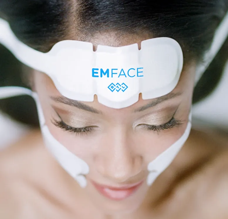 A person undergoing a cosmetic treatment with a device labeled "emface" applied to their forehead, overseen by an expert in sexual health.