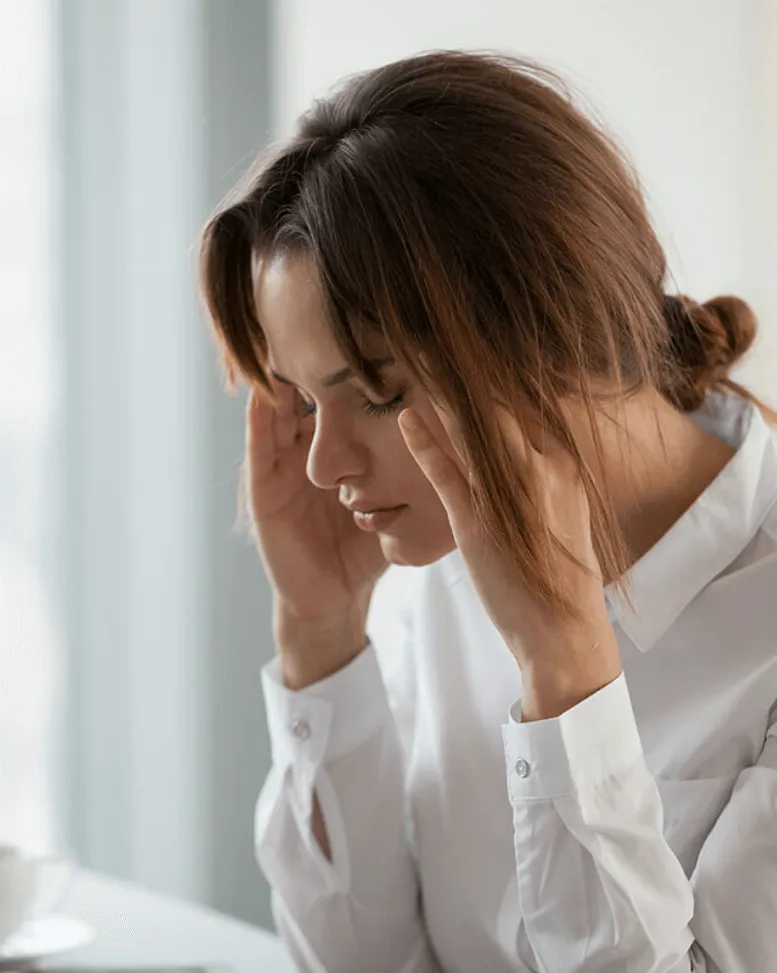 A Sexual Health Expert in Los Angeles, in a white shirt looking down with a pensive expression, her hands resting on her temples, possibly indicating stress or deep thought.